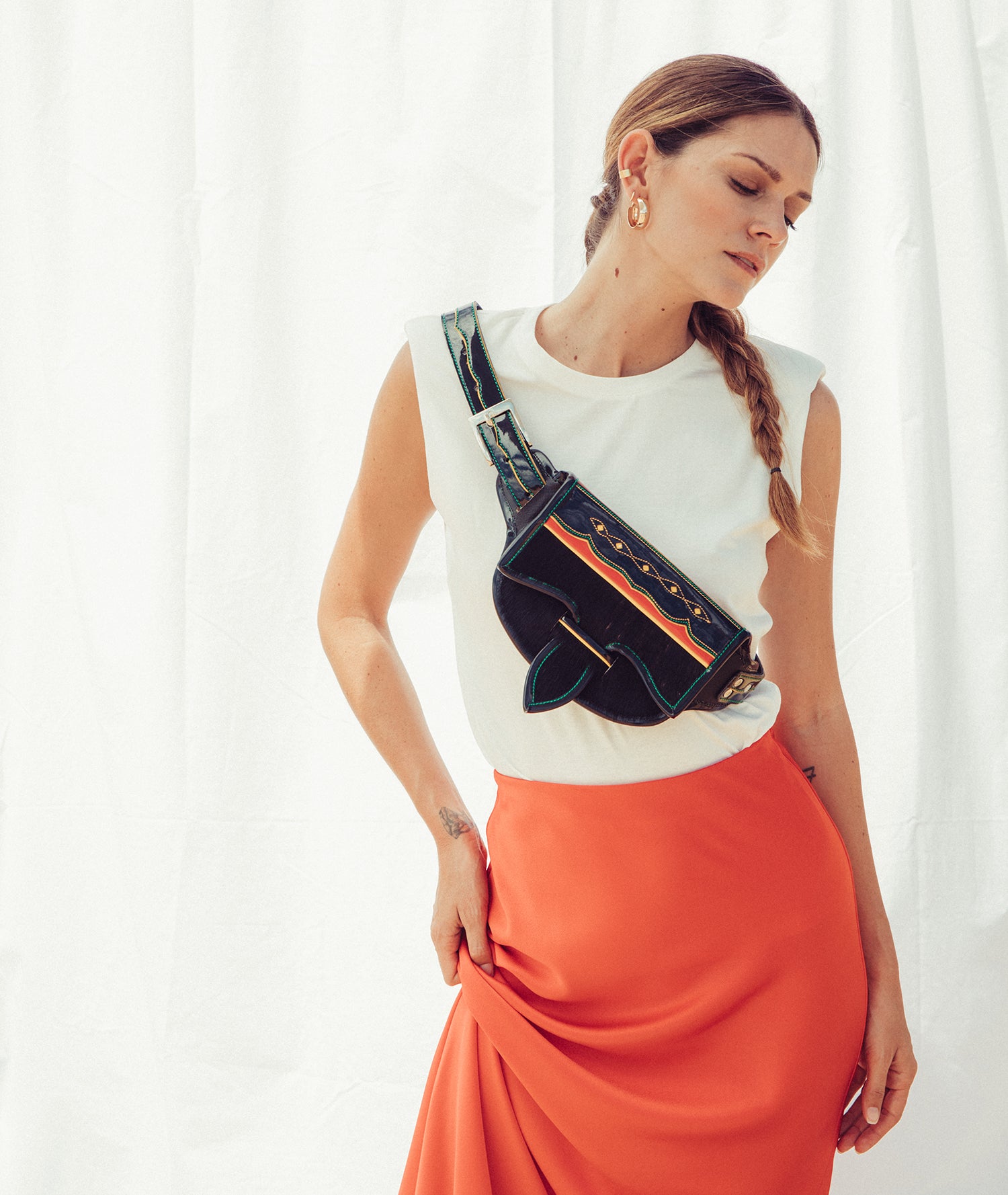 Woman wearing a white top and orange skirt with a black and blue bag over her shoulder against a white curtain background wearing a leather cowhide belt bag.