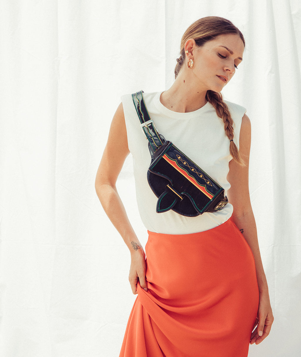 Woman wearing a white top and orange skirt with a black and blue bag over her shoulder against a white curtain background wearing a leather cowhide belt bag. 